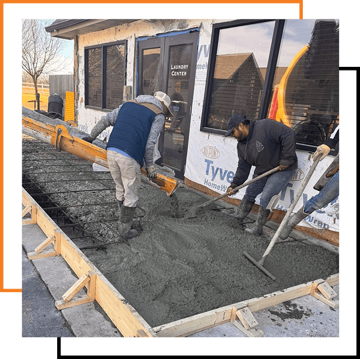 Workers pour and spread wet concrete over a rebar grid beside a building using a chute and rakes to level the surface.
