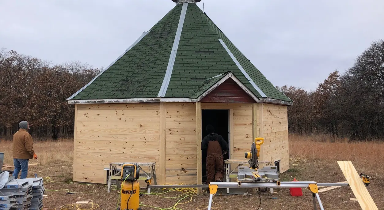 Two workers building a small wooden gazebo structure with a green shingle roof in an open grassy field, with a miter saw, power tools, lumber, and construction materials set up around the entrance.