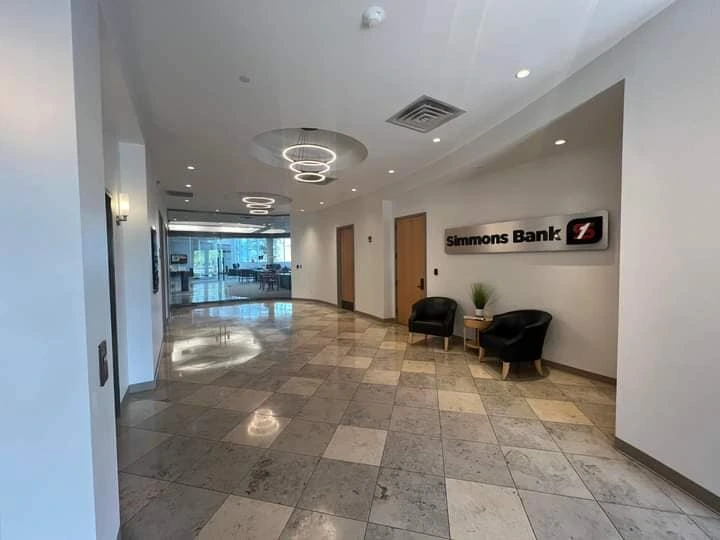 Modern bank lobby hallway with tiled flooring, circular ceiling lights, a Simmons Bank wall sign, and two black chairs with a small table near the entrance leading to a glass-walled office area.
